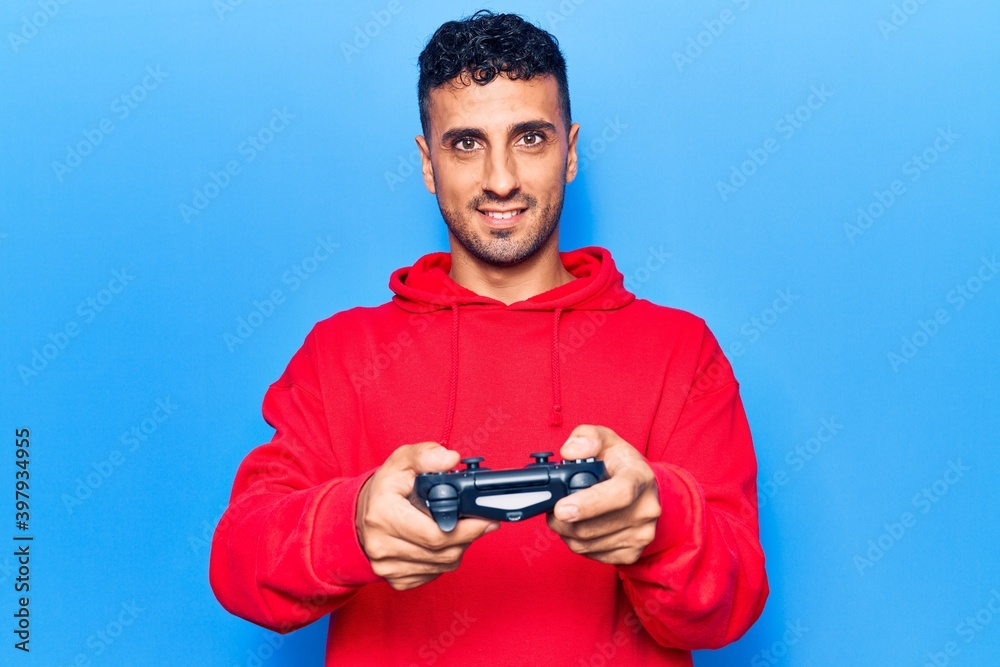 Young hispanic man playing video game holding controller looking positive and happy standing and smiling with a confident smile showing teeth