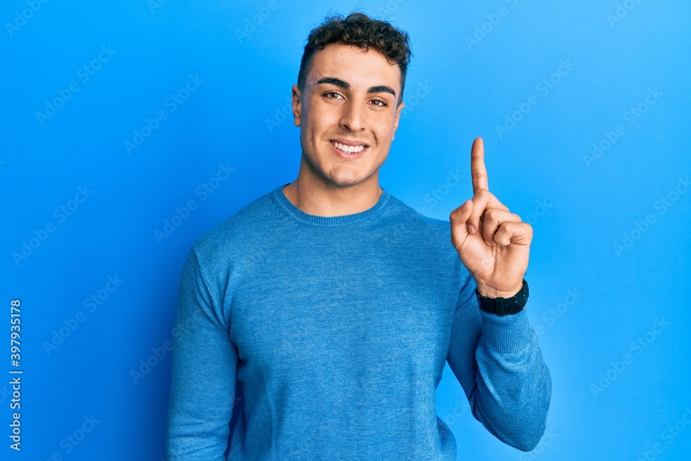 Fototapeta premium Hispanic young man wearing casual winter sweater showing and pointing up with finger number one while smiling confident and happy.