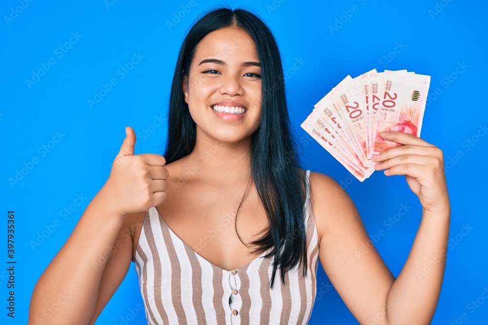 Young beautiful asian girl holding israel shekels smiling happy and positive, thumb up doing excellent and approval sign Young beautiful asian girl holding israel shekels smiling happy and positive, thumb up doing excellent and approval sign