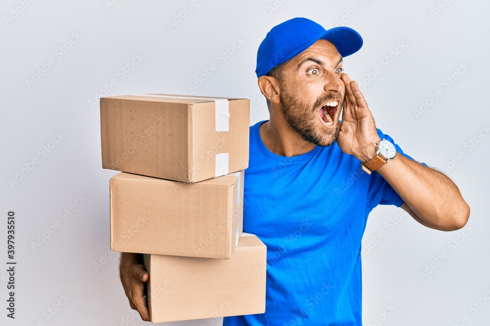 Handsome man with beard wearing courier uniform holding delivery packages shouting and screaming loud to side with hand on mouth. communication concept. Handsome man with beard wearing courier uniform holding delivery packages shouting and screaming loud to side with hand on mouth. communication concept.