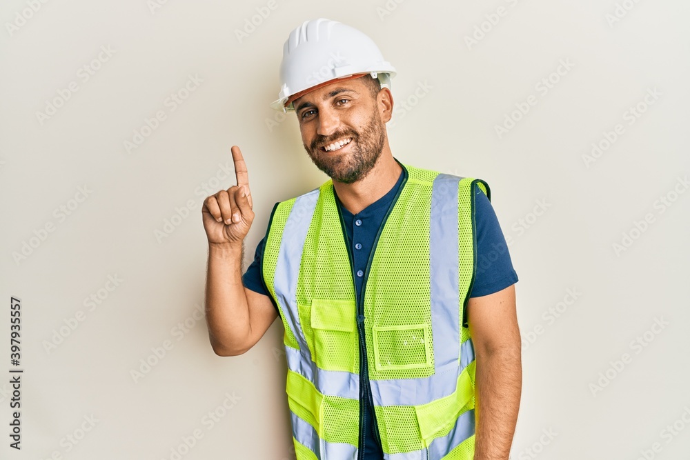 Handsome man with beard wearing safety helmet and reflective jacket smiling with an idea or question pointing finger up with happy face, number one