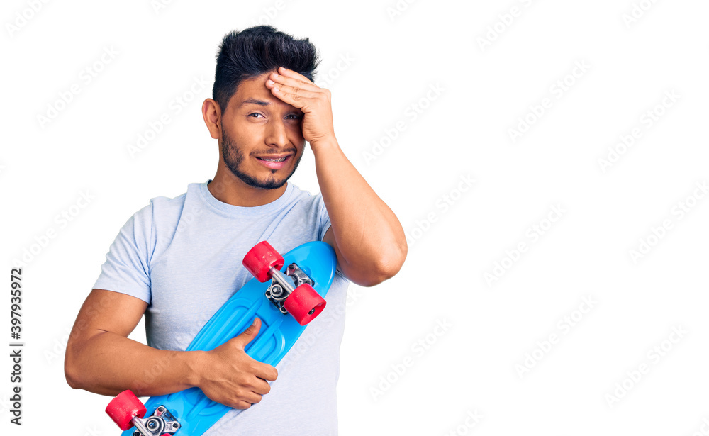 Handsome latin american young man holding skate stressed and frustrated with hand on head, surprised and angry face Handsome latin american young man holding skate stressed and frustrated with hand on head, surprised and angry face