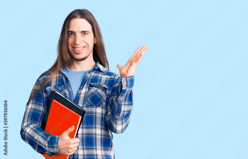 Young adult man with long hair holding collage book celebrating victory with happy smile and winner expression with raised hands Young adult man with long hair holding collage book celebrating victory with happy smile and winner expression with raised hands