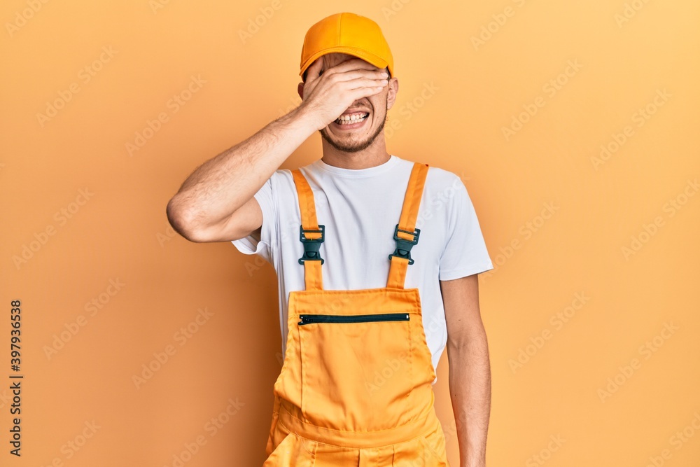 Hispanic young man wearing handyman uniform smiling and laughing with hand on face covering eyes for surprise. blind concept.