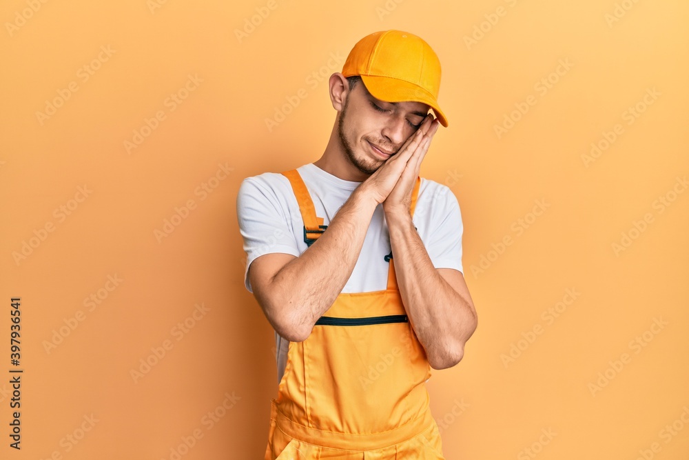 Hispanic young man wearing handyman uniform sleeping tired dreaming and posing with hands together while smiling with closed eyes.