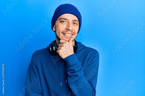 Hispanic young man wearing sweatshirt and headphones looking confident at the camera with smile with crossed arms and hand raised on chin. thinking positive.
