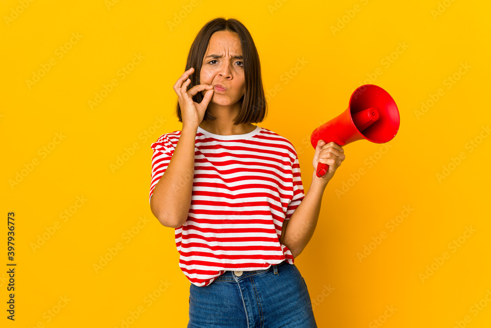 Young hispanic woman holding a megaphone with fingers on lips keeping a secret. Young hispanic woman holding a megaphone with fingers on lips keeping a secret.