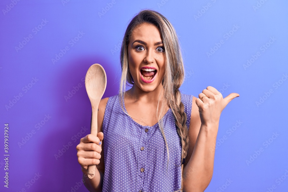 Young beautiful blonde woman holding wooden spoon over isolated purple background pointing thumb up to the side smiling happy with open mouth Young beautiful blonde woman holding wooden spoon over isolated purple background pointing thumb up to the side smiling happy with open mouth