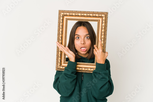 Young hispanic woman holding an old frame surprised and shocked. © Asier