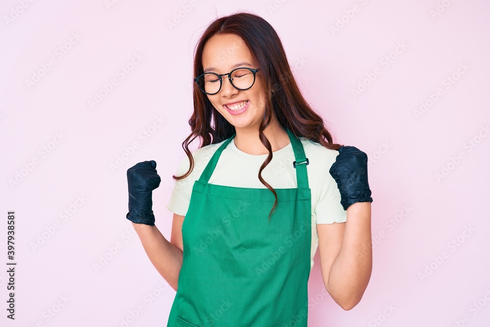 Young beautiful chinese girl wearing cleaner apron excited for success with arms raised and eyes closed celebrating victory smiling. winner concept. Young beautiful chinese girl wearing cleaner apron excited for success with arms raised and eyes closed celebrating victory smiling. winner concept.