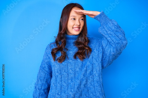 Young beautiful chinese girl wearing casual winter sweater very happy and smiling looking far away with hand over head. searching concept.