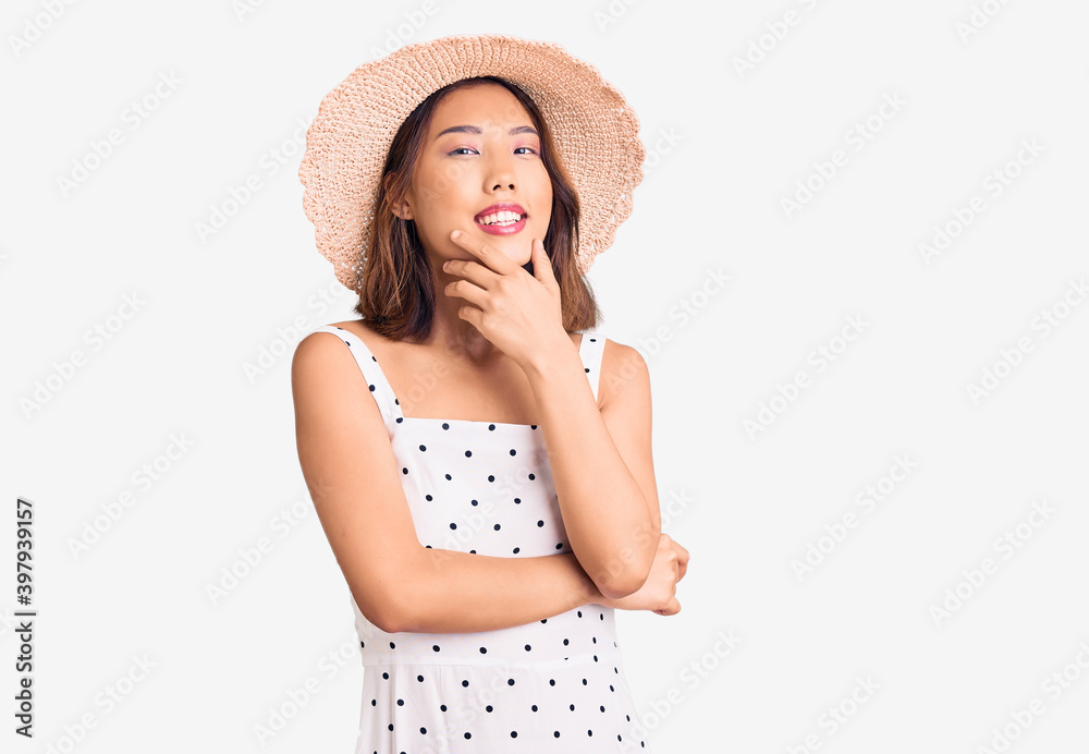 Young beautiful chinese girl wearing summer hat looking confident at the camera smiling with crossed arms and hand raised on chin. thinking positive. Young beautiful chinese girl wearing summer hat looking confident at the camera smiling with crossed arms and hand raised on chin. thinking positive.