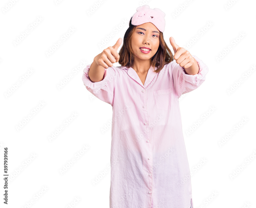 Young beautiful chinese girl wearing sleep mask and pajama approving doing positive gesture with hand, thumbs up smiling and happy for success. winner gesture. Young beautiful chinese girl wearing sleep mask and pajama approving doing positive gesture with hand, thumbs up smiling and happy for success. winner gesture.