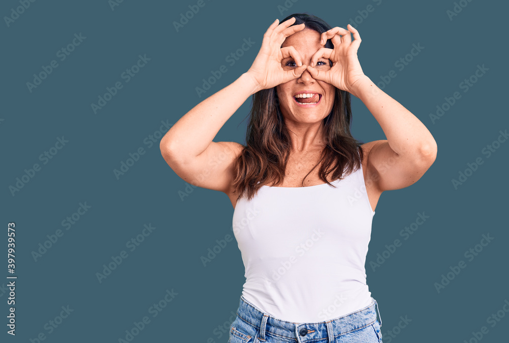 Young beautiful brunette woman wearing casual sleeveless t-shirt doing ok gesture like binoculars sticking tongue out, eyes looking through fingers. crazy expression.