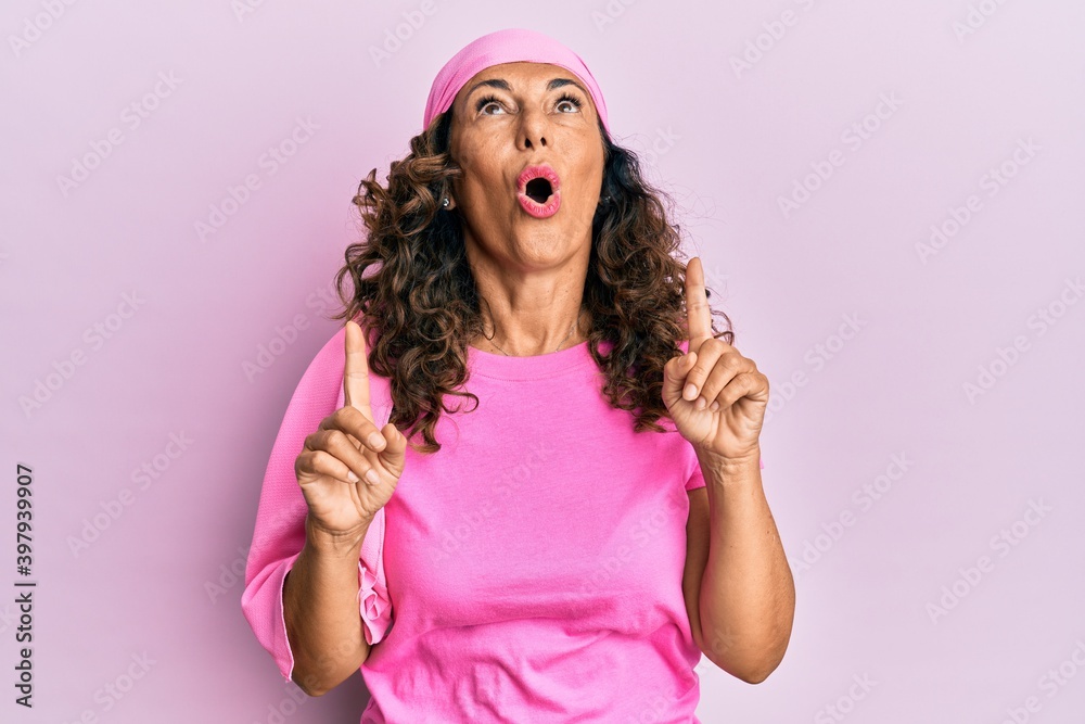 Middle age hispanic woman wearing breast cancer support pink scarf amazed and surprised looking up and pointing with fingers and raised arms. Middle age hispanic woman wearing breast cancer support pink scarf amazed and surprised looking up and pointing with fingers and raised arms.