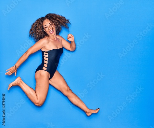 Middle age beautiful hispanic woman on vacation wearing swimsuit smiling happy. Jumping with smile on face celebrating over isolated blue background