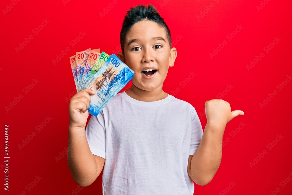 Little boy hispanic kid holding swiss franc banknotes pointing thumb up to the side smiling happy with open mouth Little boy hispanic kid holding swiss franc banknotes pointing thumb up to the side smiling happy with open mouth