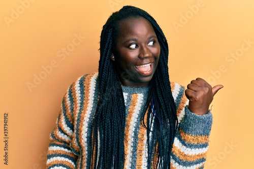 Young black woman with braids wearing casual winter sweater smiling with happy face looking and pointing to the side with thumb up. © Krakenimages.com