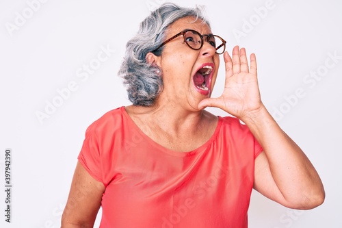 Senior hispanic grey- haired woman wearing casual clothes and glasses shouting and screaming loud to side with hand on mouth. communication concept.