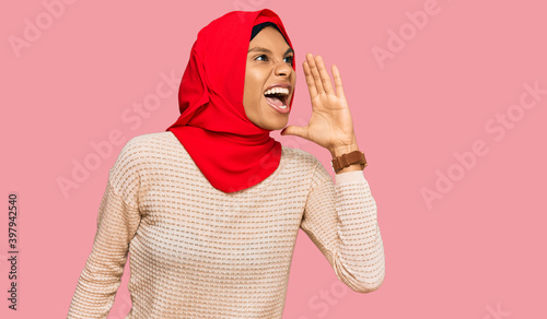 Young african american woman wearing traditional islamic hijab scarf shouting and screaming loud to side with hand on mouth. communication concept.