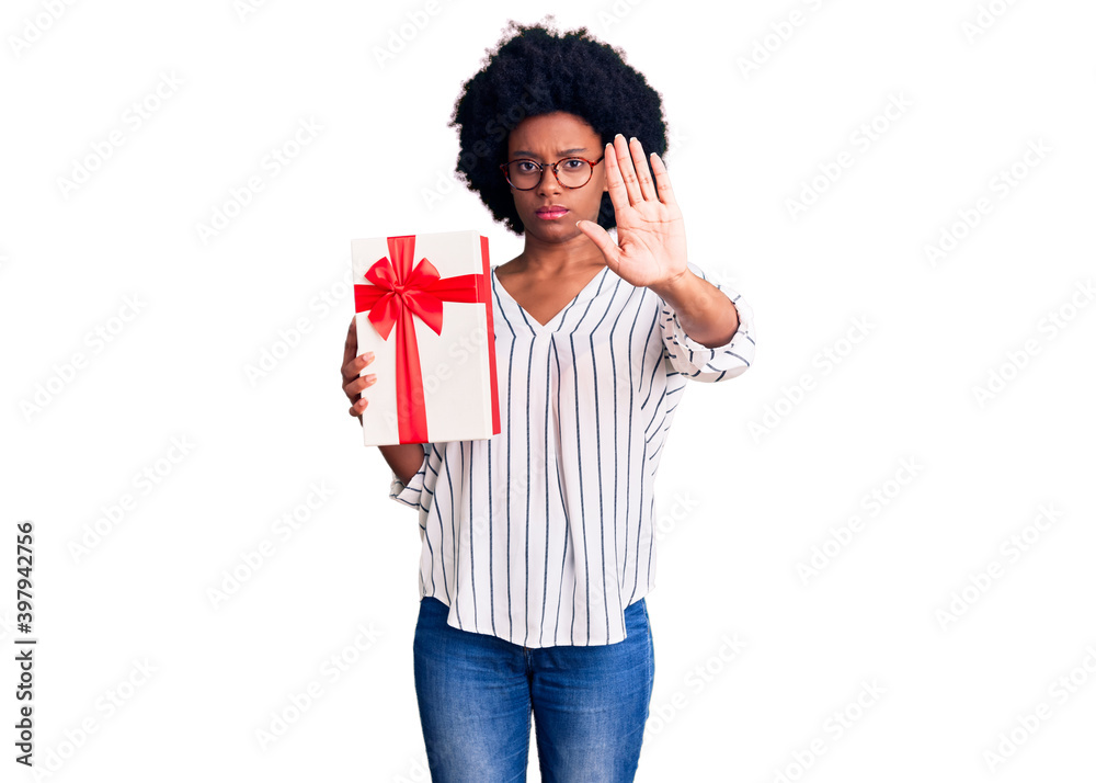 Young african american woman holding gift with open hand doing stop sign with serious and confident expression, defense gesture Young african american woman holding gift with open hand doing stop sign with serious and confident expression, defense gesture