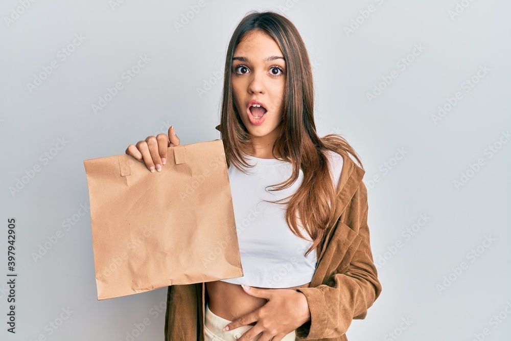 Young brunette woman holding take away paper bag scared and amazed with open mouth for surprise, disbelief face Young brunette woman holding take away paper bag scared and amazed with open mouth for surprise, disbelief face