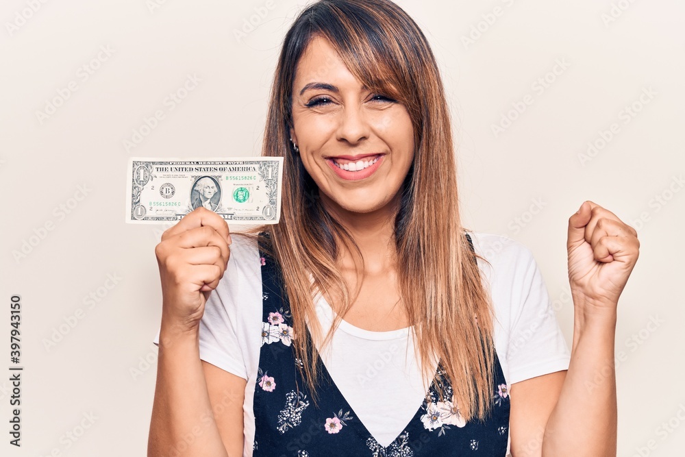 Young beautiful woman holding one dollar screaming proud, celebrating victory and success very excited with raised arm Young beautiful woman holding one dollar screaming proud, celebrating victory and success very excited with raised arm
