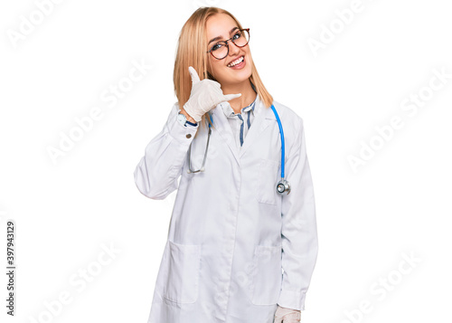 Beautiful caucasian woman wearing doctor uniform and stethoscope smiling doing phone gesture with hand and fingers like talking on the telephone. communicating concepts.