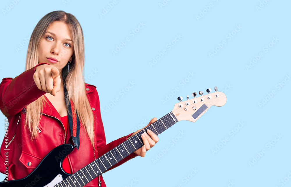 Young beautiful blonde woman playing electric guitar pointing with finger to the camera and to you, confident gesture looking serious Young beautiful blonde woman playing electric guitar pointing with finger to the camera and to you, confident gesture looking serious