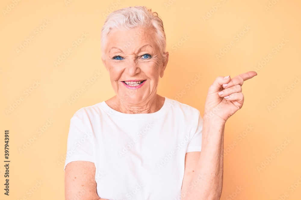Senior beautiful woman with blue eyes and grey hair wearing classic white tshirt over yellow background with a big smile on face, pointing with hand and finger to the side looking at the camera. Senior beautiful woman with blue eyes and grey hair wearing classic white tshirt over yellow background with a big smile on face, pointing with hand and finger to the side looking at the camera.