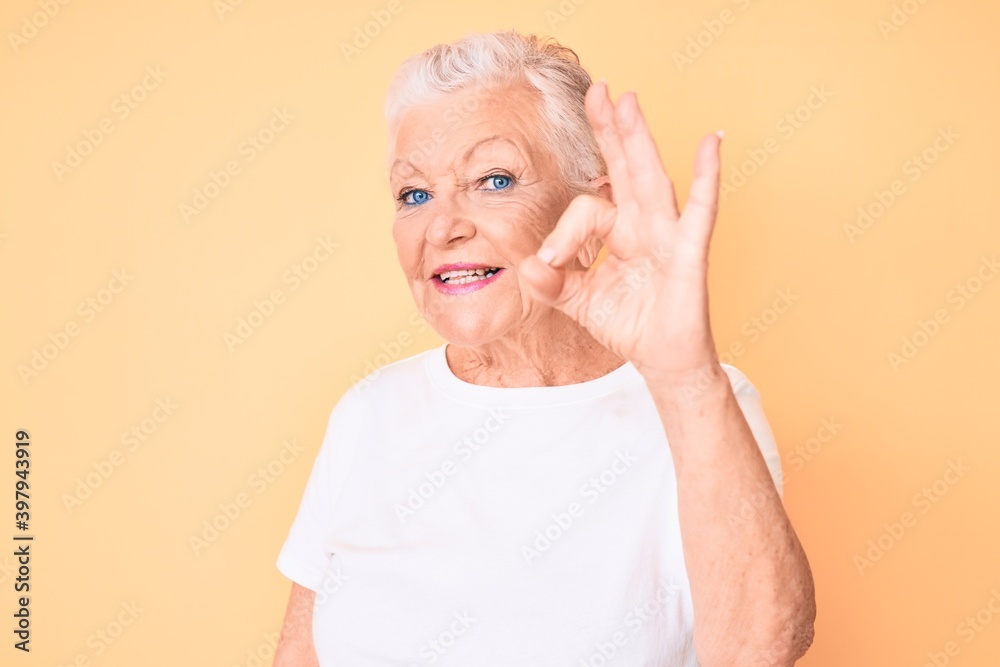 Senior beautiful woman with blue eyes and grey hair wearing classic white tshirt over yellow background smiling positive doing ok sign with hand and fingers. successful expression. Senior beautiful woman with blue eyes and grey hair wearing classic white tshirt over yellow background smiling positive doing ok sign with hand and fingers. successful expression.