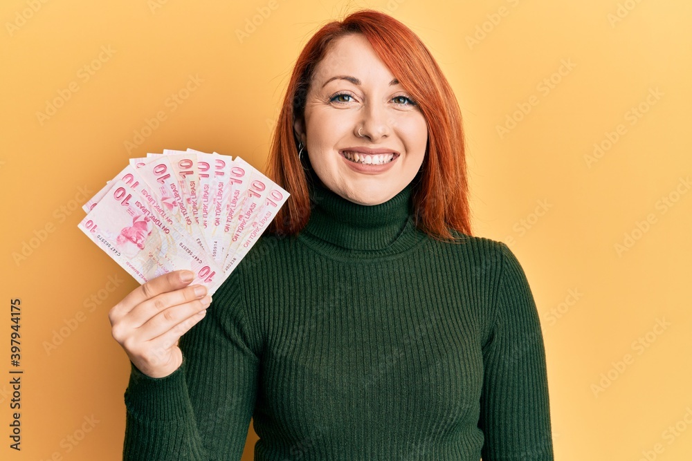 Beautiful redhead woman holding 10 turkish lira banknotes looking positive and happy standing and smiling with a confident smile showing teeth Beautiful redhead woman holding 10 turkish lira banknotes looking positive and happy standing and smiling with a confident smile showing teeth