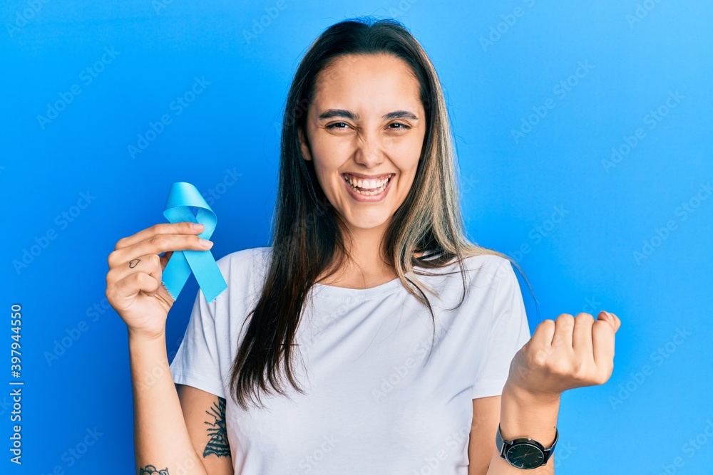 Young hispanic woman holding blue ribbon screaming proud, celebrating victory and success very excited with raised arms Young hispanic woman holding blue ribbon screaming proud, celebrating victory and success very excited with raised arms