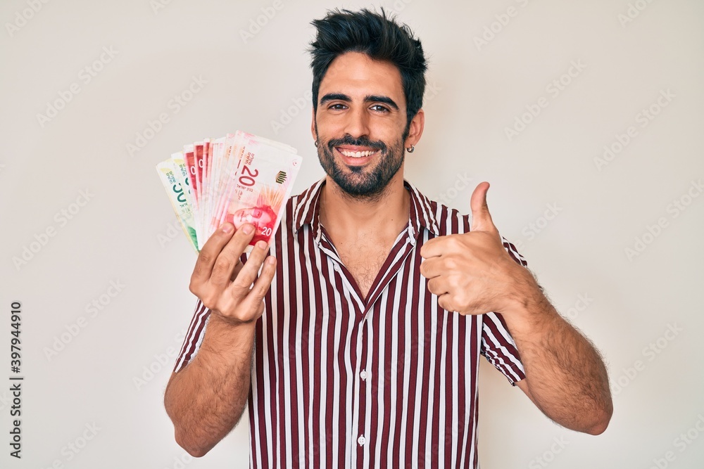 Handsome hispanic man with beard holding israel shekels smiling happy and positive, thumb up doing excellent and approval sign Handsome hispanic man with beard holding israel shekels smiling happy and positive, thumb up doing excellent and approval sign