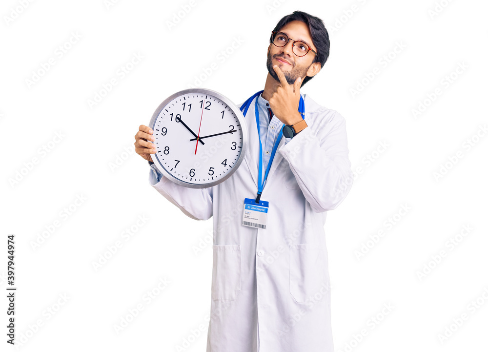 Handsome hispanic man wearing doctor uniform holding clock serious face thinking about question with hand on chin, thoughtful about confusing idea Handsome hispanic man wearing doctor uniform holding clock serious face thinking about question with hand on chin, thoughtful about confusing idea