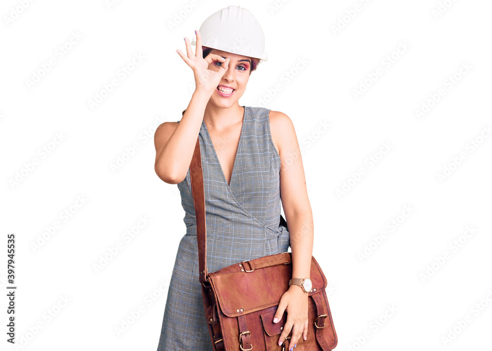 Young beautiful woman wearing architect hardhat and leather bag smiling happy doing ok sign with hand on eye looking through fingers Young beautiful woman wearing architect hardhat and leather bag smiling happy doing ok sign with hand on eye looking through fingers