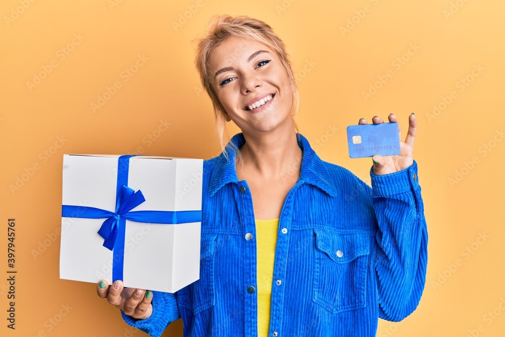 Young blonde girl holding gift and credit card smiling with a happy and cool smile on face. showing teeth. Young blonde girl holding gift and credit card smiling with a happy and cool smile on face. showing teeth.