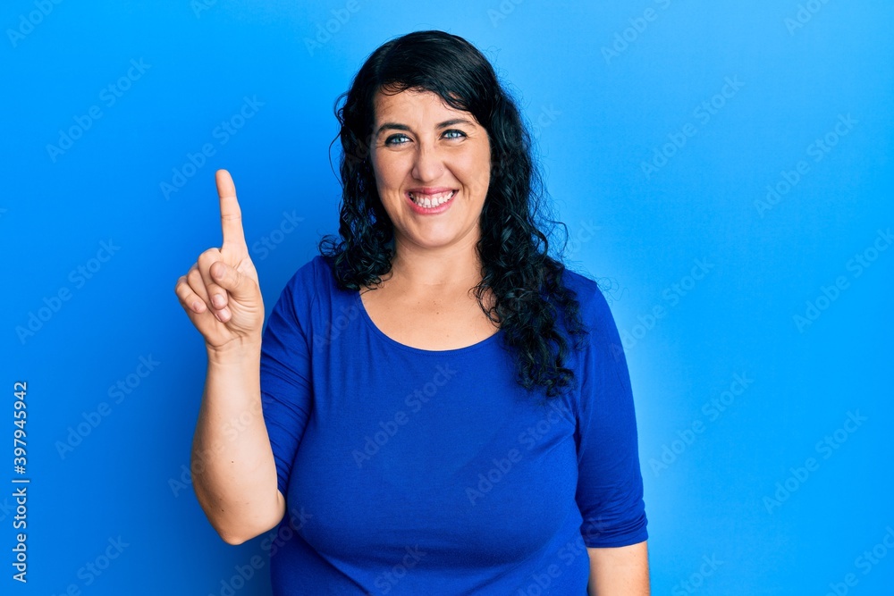 Plus size brunette woman wearing casual blue shirt showing and pointing up with finger number one while smiling confident and happy.