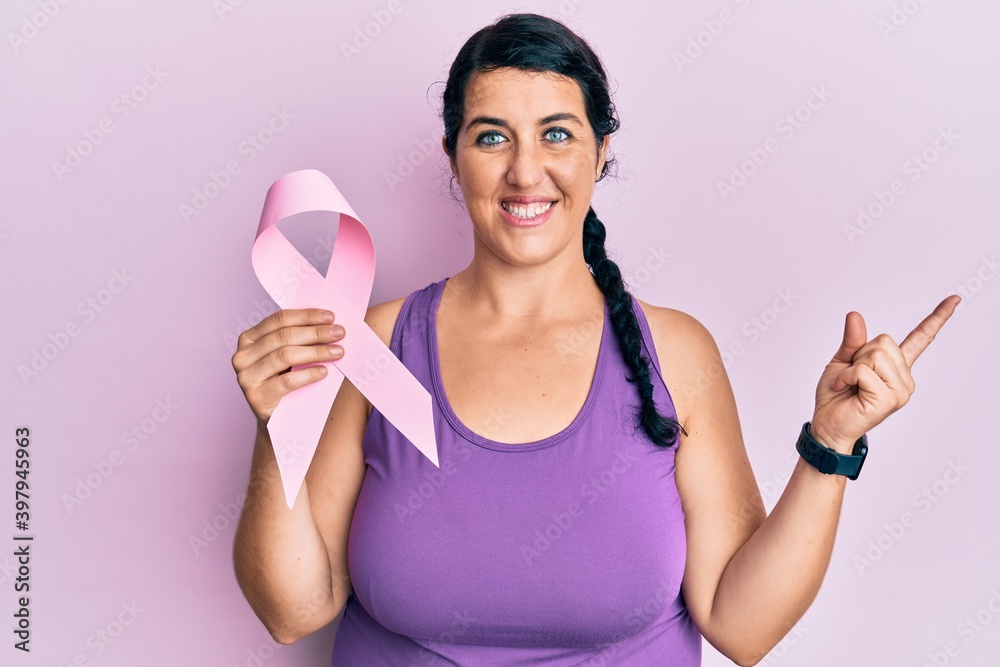 Plus size brunette woman holding pink cancer ribbon smiling happy pointing with hand and finger to the side Plus size brunette woman holding pink cancer ribbon smiling happy pointing with hand and finger to the side