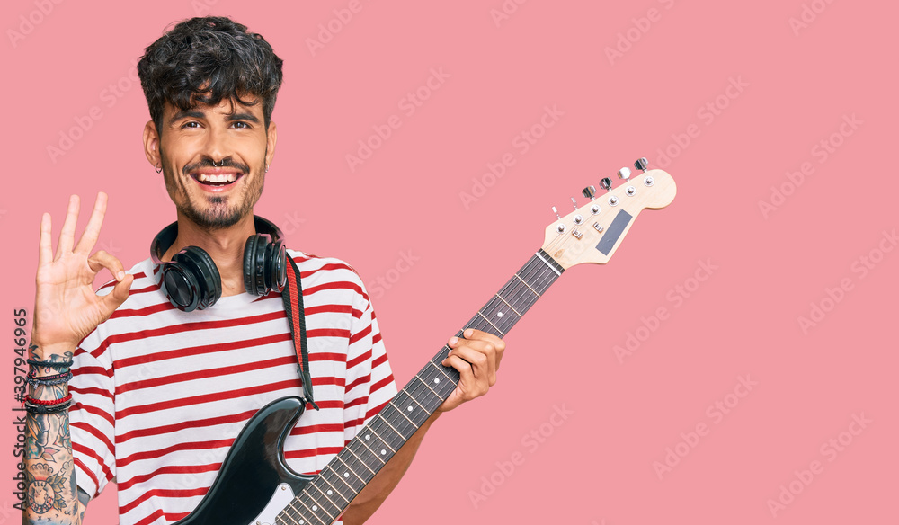 Obraz premium Young hispanic man playing electric guitar doing ok sign with fingers, smiling friendly gesturing excellent symbol