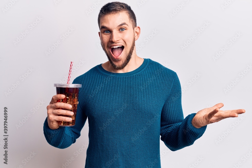 Young handsome man dirnking cola refreshment beverage celebrating achievement with happy smile and winner expression with raised hand Young handsome man dirnking cola refreshment beverage celebrating achievement with happy smile and winner expression with raised hand