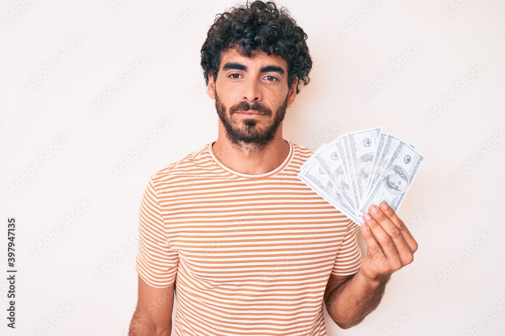 Handsome young man with curly hair and bear holding money bag with dollar symbol thinking attitude and sober expression looking self confident Handsome young man with curly hair and bear holding money bag with dollar symbol thinking attitude and sober expression looking self confident