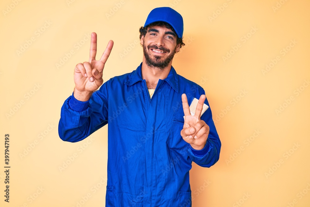 Handsome young man with curly hair and bear wearing builder jumpsuit uniform smiling looking to the camera showing fingers doing victory sign. number two.