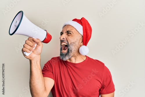Middle age grey-haired man wearing santa claus hat screaming angry using megaphone over isolated white background.