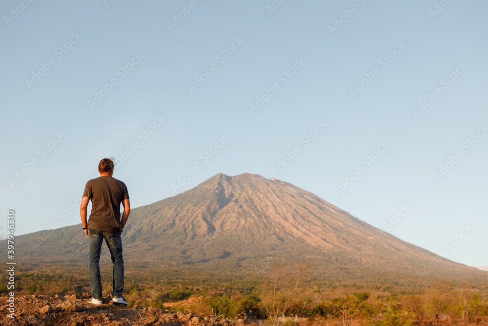 Rear view young man traveler standing on top of rock  and enjoying amazing volcanic mountain Agung view in Bali during sunrise. Travel and active lifestyle concept.