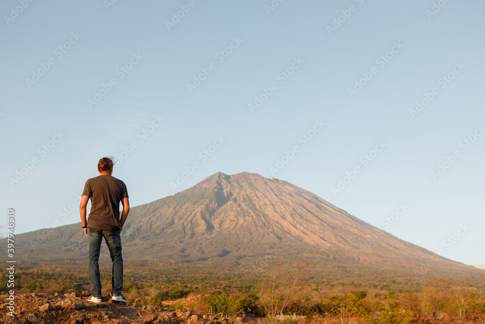 Rear view young man traveler standing on top of rock and enjoying amazing volcanic mountain Agung view in Bali during sunrise. Travel and active lifestyle concept. Rear view young man traveler standing on top of rock and enjoying amazing volcanic mountain Agung view in Bali during sunrise. Travel and active lifestyle concept.