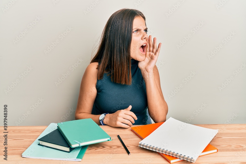 Young caucasian woman studying for exam shouting and screaming loud to side with hand on mouth. communication concept. Young caucasian woman studying for exam shouting and screaming loud to side with hand on mouth. communication concept.