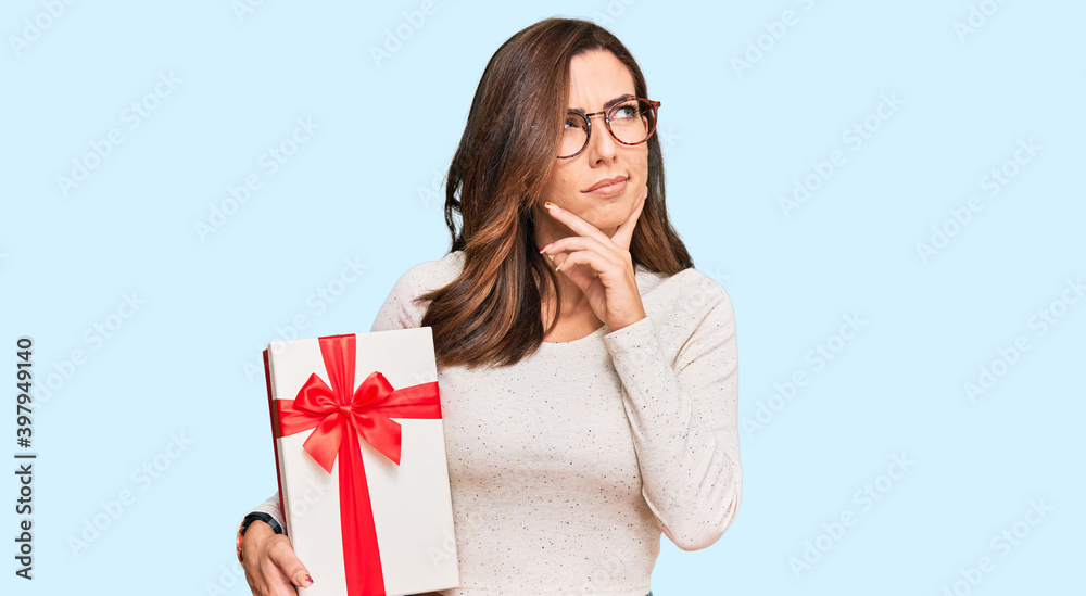Young brunette woman holding gift serious face thinking about question with hand on chin, thoughtful about confusing idea Young brunette woman holding gift serious face thinking about question with hand on chin, thoughtful about confusing idea