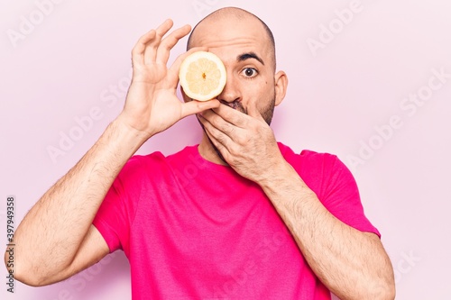 Young handsome bald man holding slice of lemon over eye covering mouth with hand, shocked and afraid for mistake. surprised expression