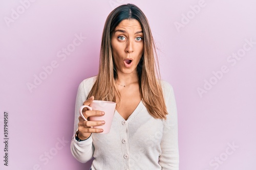 Brunette young woman drinking a cup of coffee scared and amazed with open mouth for surprise, disbelief face
